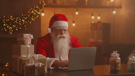 Weihnachtsmann-Benutzt-Laptop-Im-Wohnzimmer.-Porträt-Des-Nachdenklichen-Weihnachtsmanns,-Der-Mit-Modernem-Laptop-In-Dekoriertem-Haus-Tippt.-Hochwertiges-4K-Filmmaterial