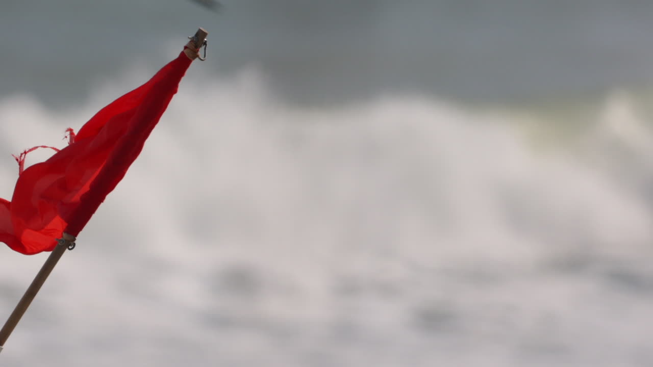 Primo piano ravvicinato di una bandiera rossa mossa dal vento marino, con le onde che si infrangono sullo sfondo