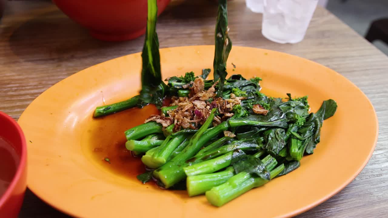 A hand uses red chopsticks to pick up Chinese broccoli from an orange plate in a casual dining setting with bright, even lighting and a close-up perspective