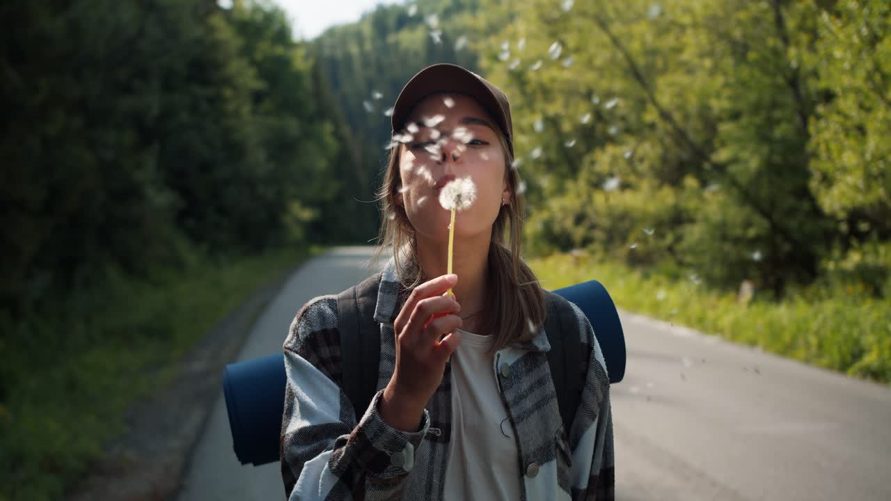 Portrait of a blonde girl who is wearing specially hiking clothes blowing on a dandelion that scatters. Happy girl tourist