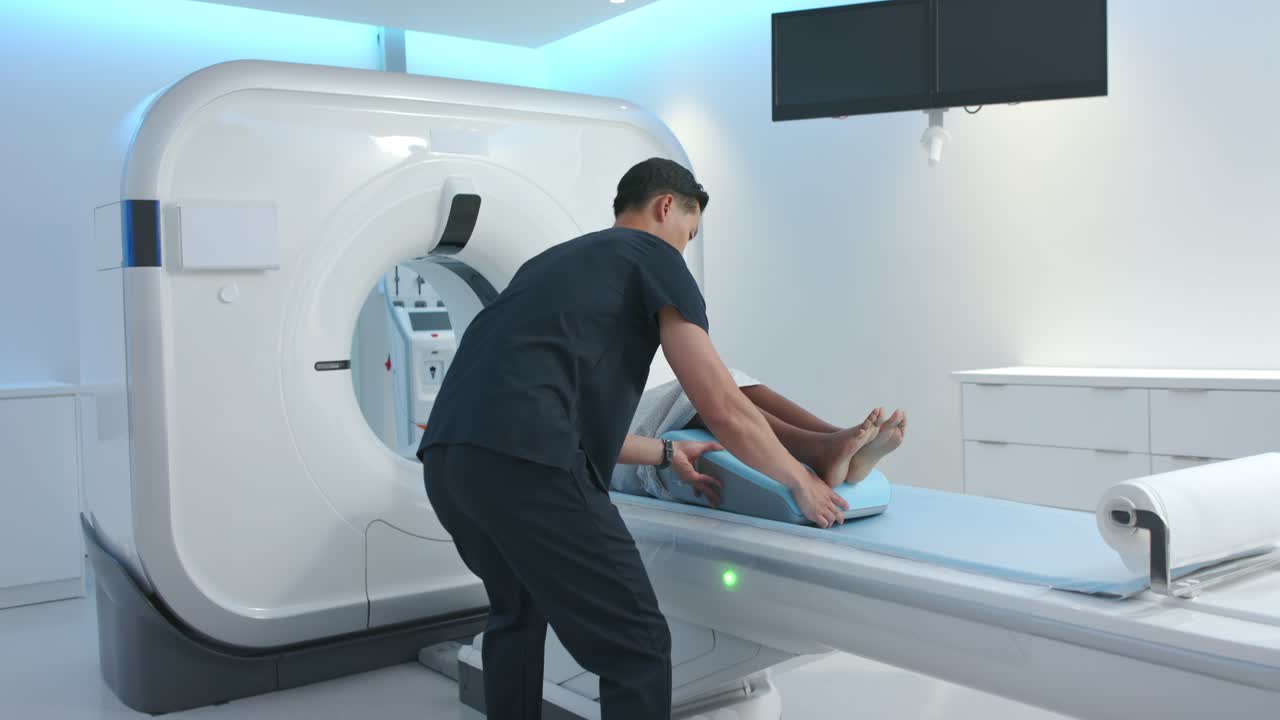 Technologist is grabbing light-blue leg wedge and is sliding it under patient's calves for CT scan