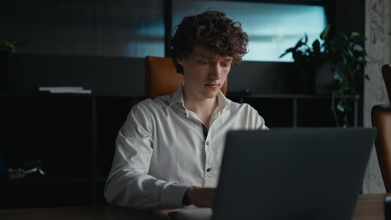 사무실에서 일하는 동안 회색 노트북에서 타이핑하는  셔츠를 입은 곱슬 머리카락을 가진 자신감있는 젊은 남자