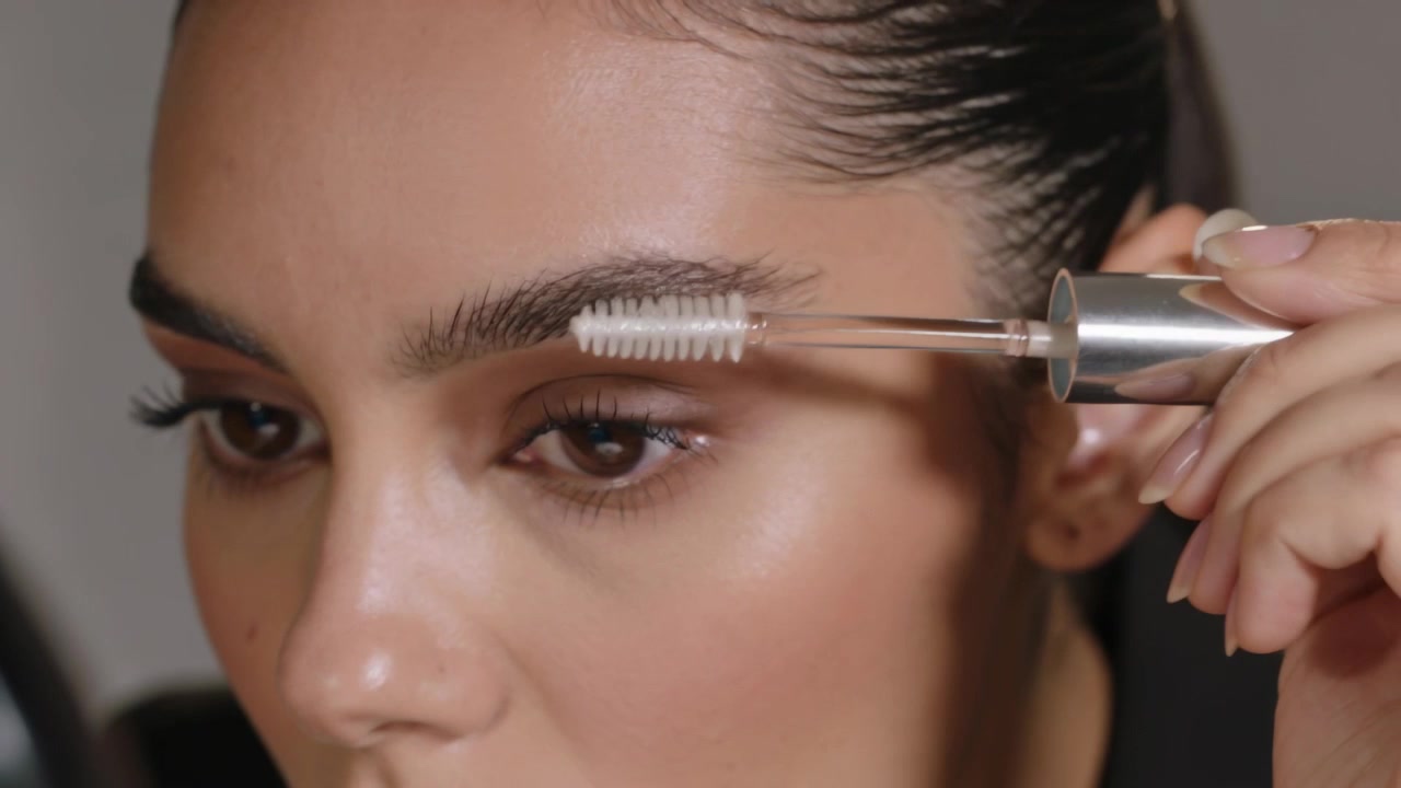 Brushing woman grooming right brow in makeup studio, spoolie wand approaching inner brow shaping