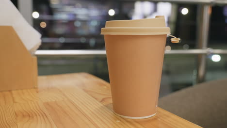 close up brown takeaway coffee cup with lid placed on wooden table in cozy restaurant interior during evening time with blurred background lights creating warm ambient atmosphere