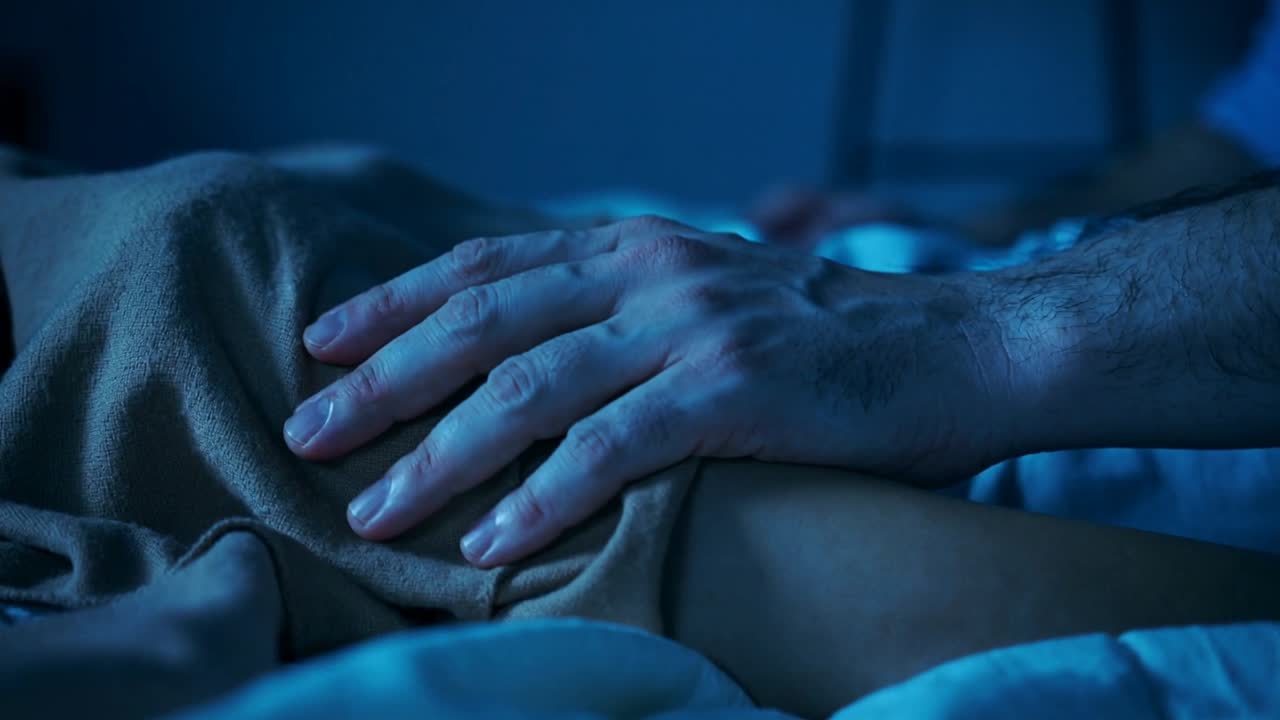 Avambraccio di un uomo in maglietta a maniche corte che preme e appoggia il palmo su una coperta marrone in camera da letto di notte.