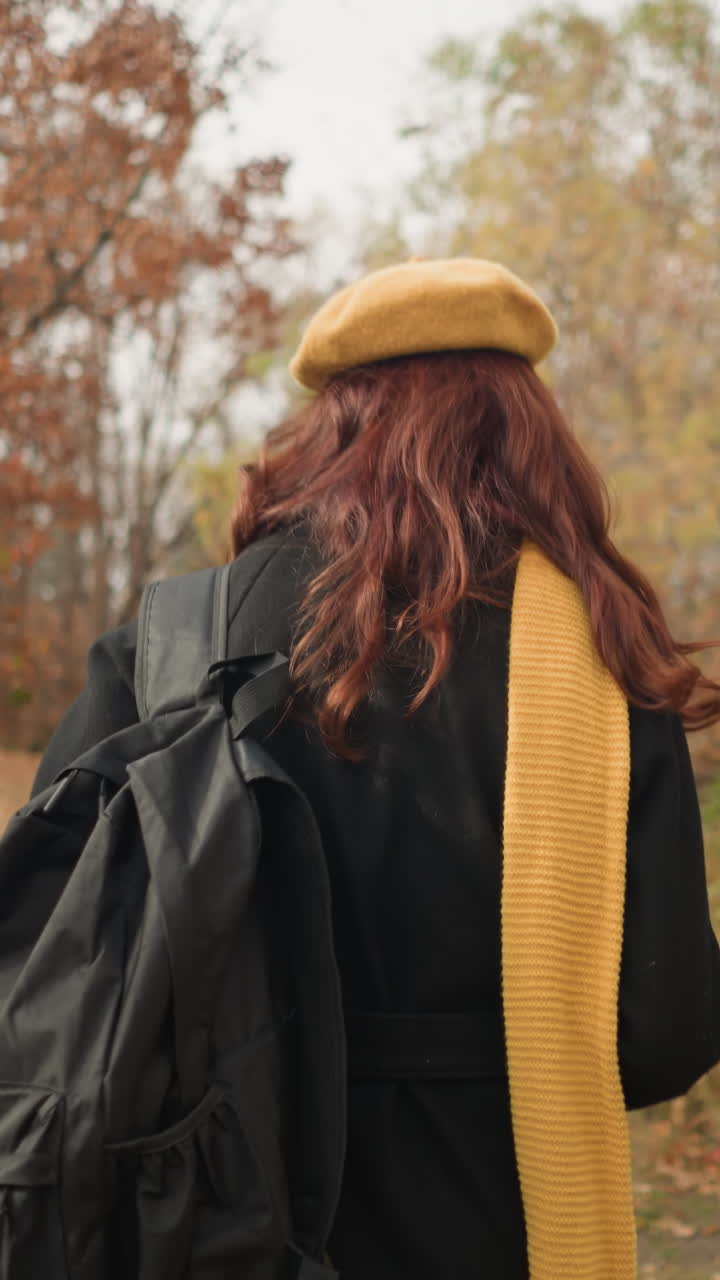 노란색 베레트, 노란색 머플러, 검은색 가방을 입은 여자가 황금색과 빨간색 잎으로 가득 찬 조용한 가을 숲을 가로질러 휘어지는 길을 걸어다니고 있습니다.