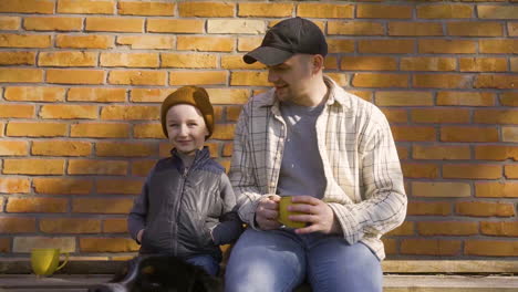 Caucasian-man-holding-a-cup-of-tea-sitting-with-her-son-outside-a-country-house-and-smiling-at-camera