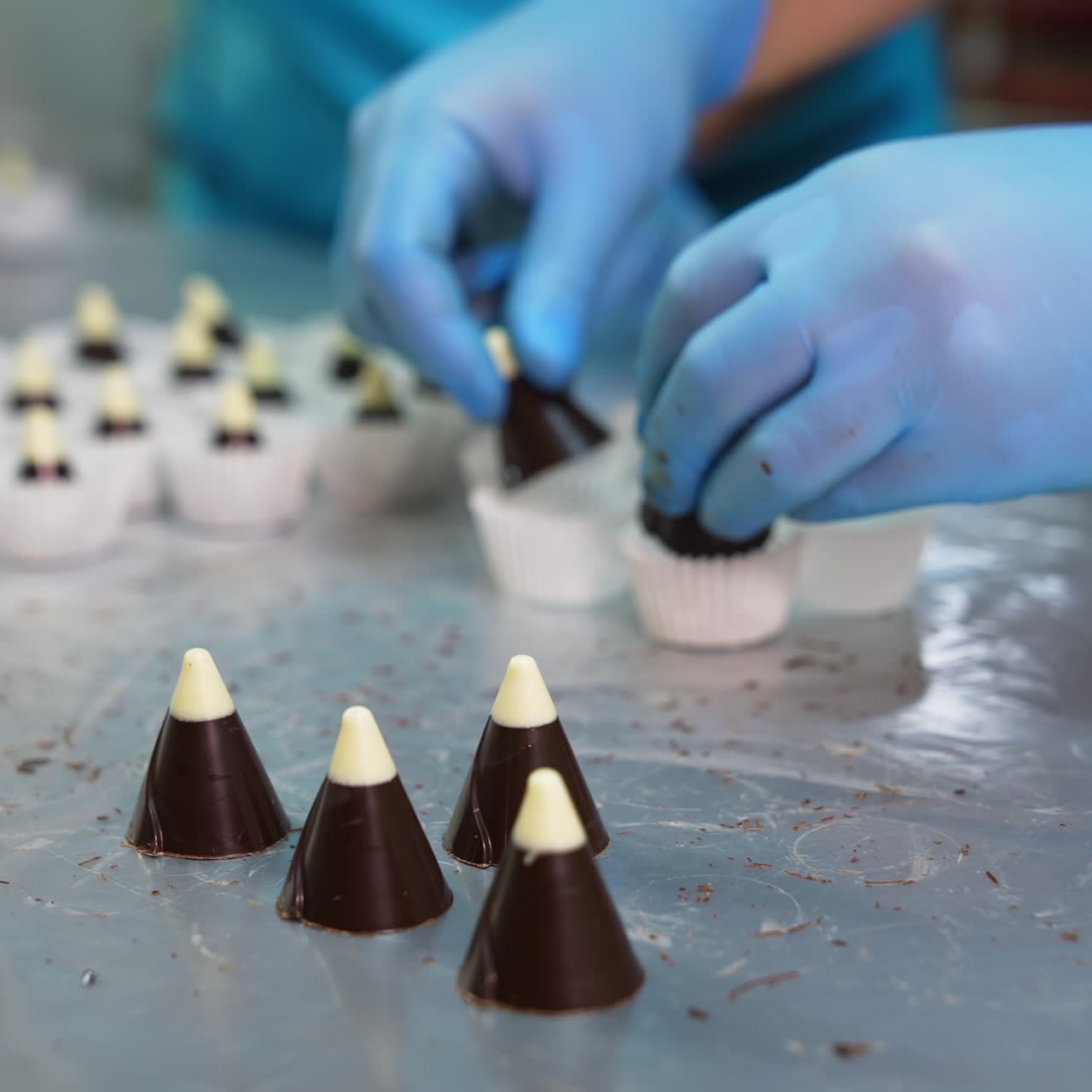 Confectioner takes little chocolate cones with white tops and puts them in paper. Packaging ready-made sweets at factory. Blurred backdrop