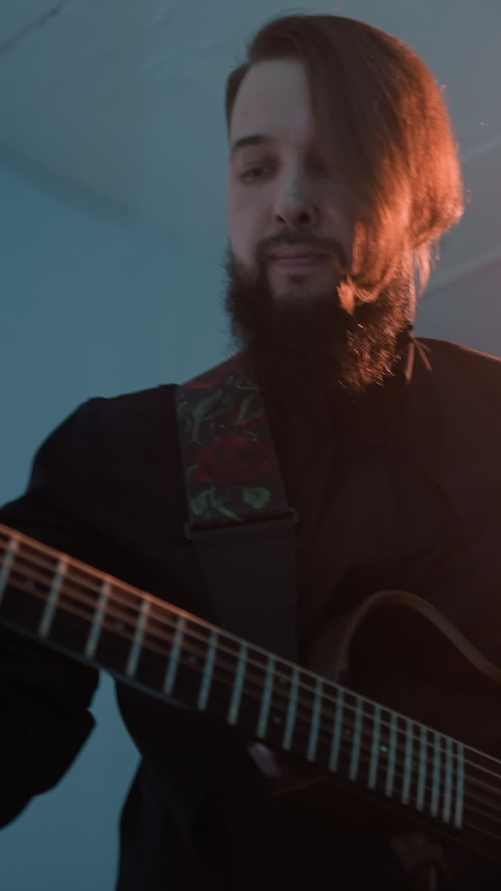 A guitarist in a black outfit, standing and playing a red electric guitar against a blue background with a bright light in the background. The musician appears deeply focused on the performance