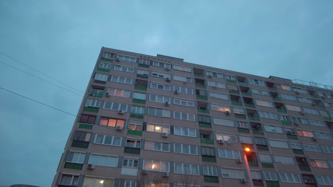 Perspectiva cinematográfica ascendente giratoria de una enorme fachada de edificio residencial en Budapest. Un cable eléctrico diagonal cruza el cielo crepuscular sobre los patrones repetitivos de balcones del bloque de viviendas