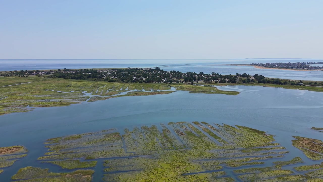 Insenature tidali serpeggianti si snodano attraverso l'erba verde della palude salmastra sotto un cielo azzurro velato, una lontana spiaggia sabbiosa e case costiere fiancheggiano la costa vicino a Salisbury Beach, Massachusetts. Ripresa aerea con drone.