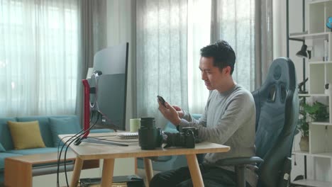 asian cameraman in long sleeved t-shirt and black pants using mobile phone while using desktop computer for working at home.