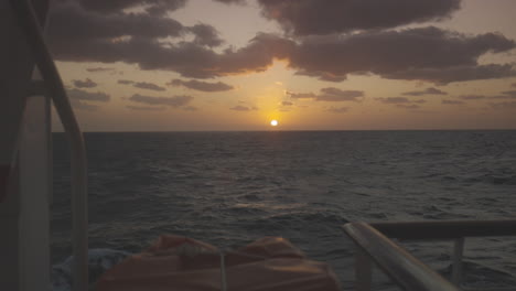 sunset at the horizon in the ocean from a deck on a slowly rooling vessel on a cloudy day. wide trucking shot
