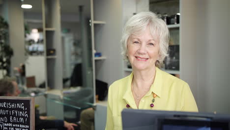 Portrait-Of-Senior-Female-Receptionist-Or-Business-Owner-At-Front-Desk-In-Hairdressing-Salon