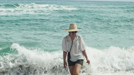 Mujer-Pasea-Por-La-Playa-Mientras-Las-Olas-De-Verano-Salpican-Sus-Piernas,-Cámara-Lenta