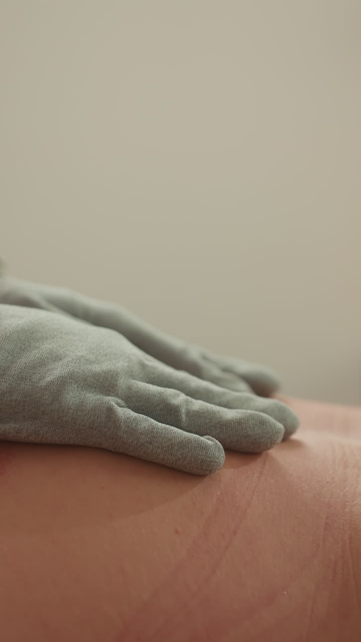 Cliente caucasica che riceve un massaggio con guanti a microcorrente in una stanza spa poco illuminata, estetista usa guanti conduttivi e clip elettrodo sulla schiena nuda producendo una breve impronta rossa, focus sul rilascio della tensione muscolare.