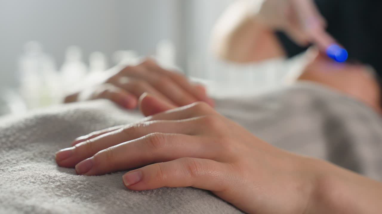 Close up of female client hands resting under blanket on clinical bed with therapist using ultrasound massage tool under blue LED light in soft focus background during spa treatment session