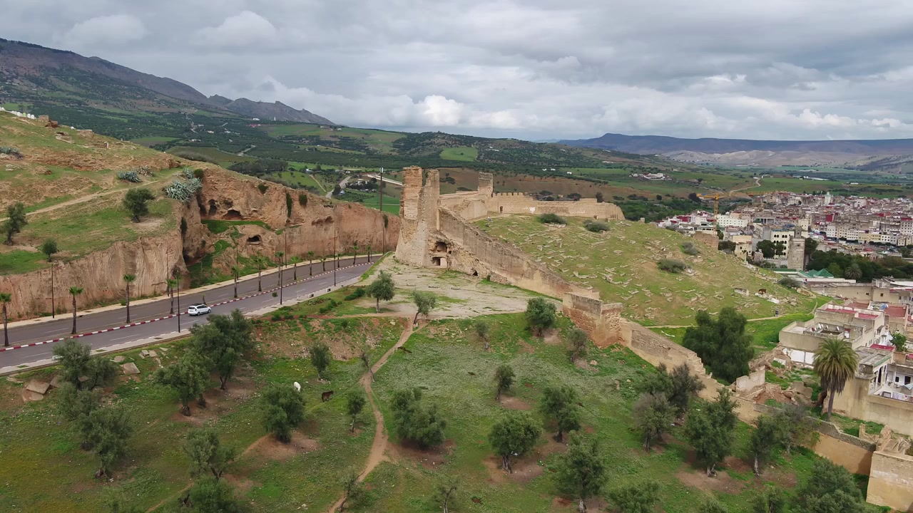 Ampia vista aerea delle storiche mura fortificate di Fez, Marocco, con una torre difensiva prominente, bastioni collinari, una strada moderna e la campagna verde e le montagne circostanti.