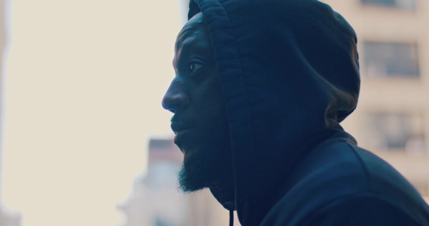 New York, USA, 21 May 2023: Man in Dark Hoodie Side Profile in Low Light Urban Setting. A Black man in a dark navy hoodie appears in side profile with sided lighting against a blurred urban background