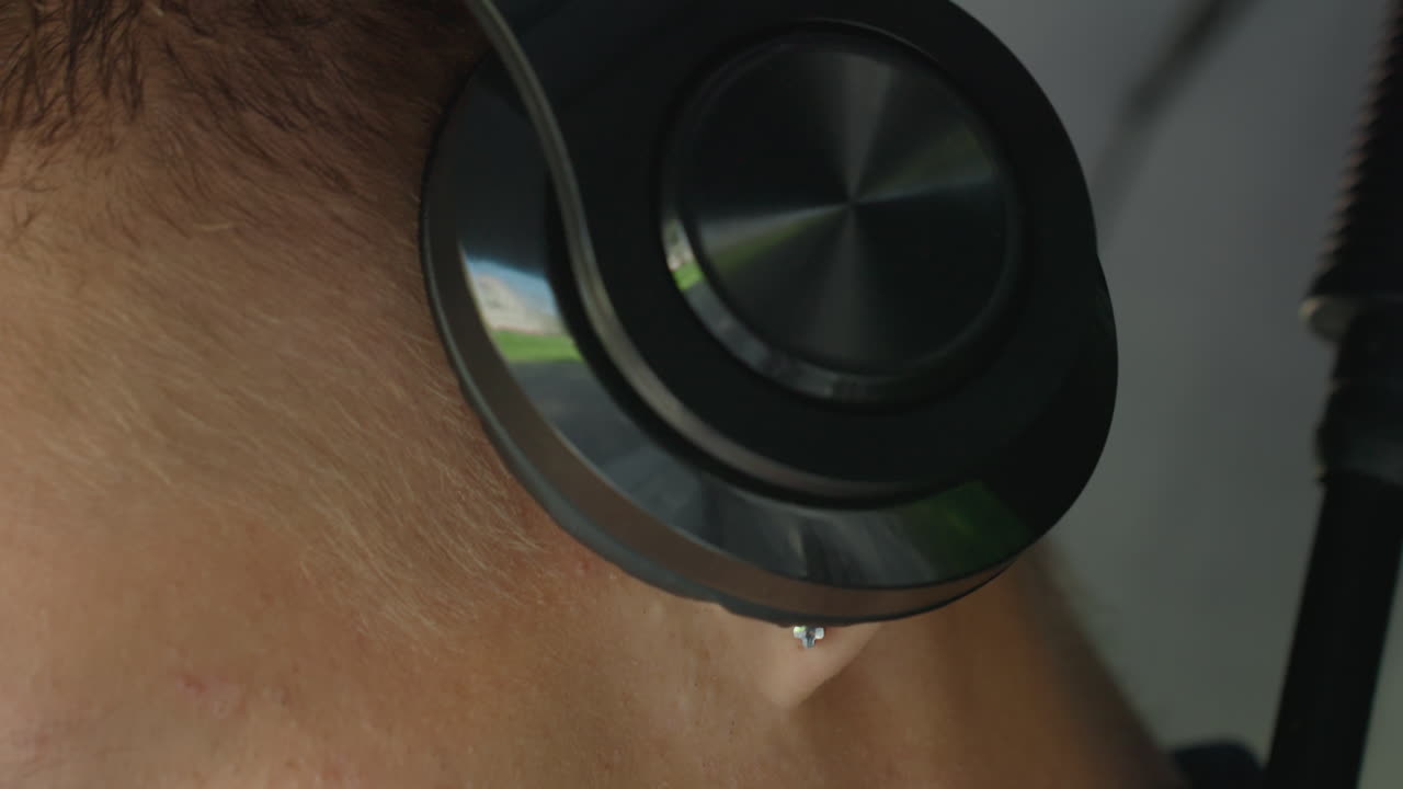 Ekstremt nærbillede af en hvid mands øre med sorte hovedtelefoner. En stud-piercing er synlig, og den reflekterende driver fanger blødt studielys. Intim lyttestemning, lyddesigner overvåger samples, fokus