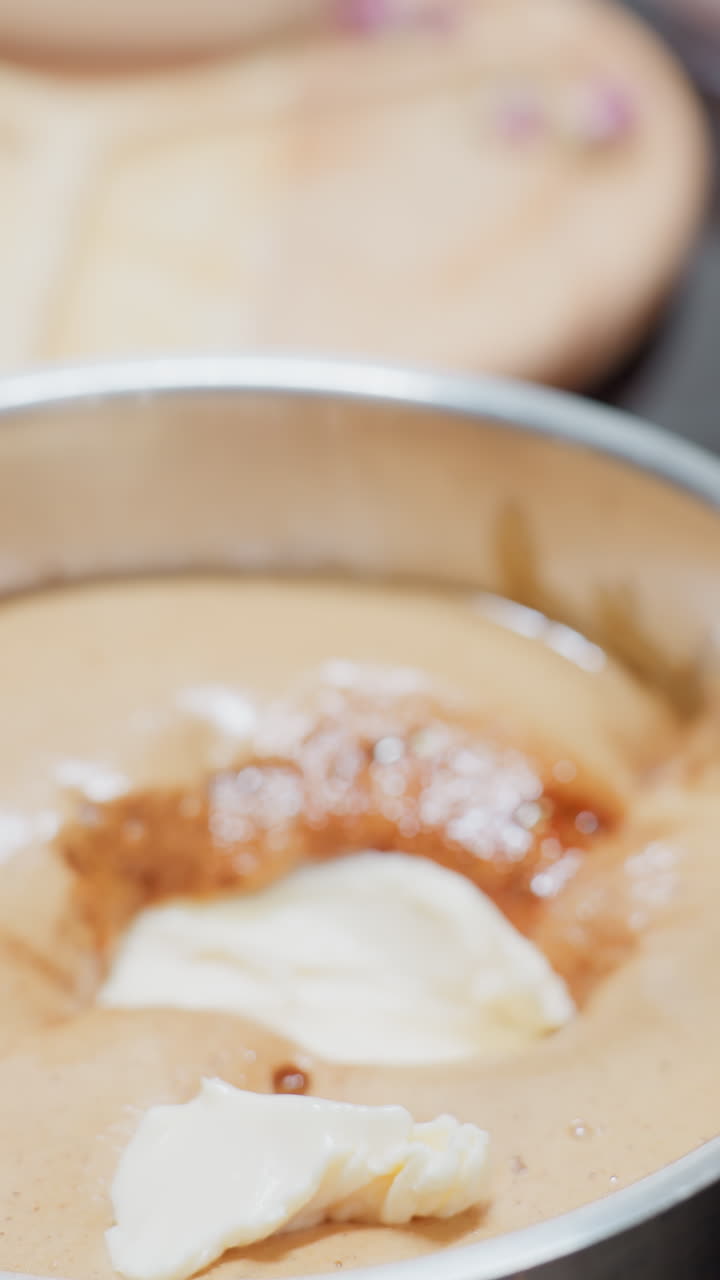 vista de cerca de la mano de una persona que sostiene mantequilla cremosa en la punta del dedo, preparándose para dejarla caer en una olla de papel marrón liso en una cocina brillante