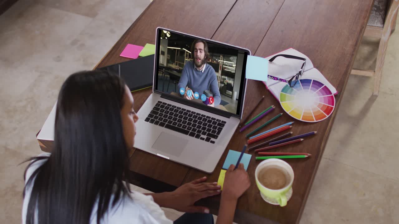 Mujer caucásica usando una computadora portátil en una videollamada con un colega y tomando notas.