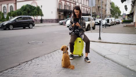 woman with her dog sitting on a suitcase on a city street.