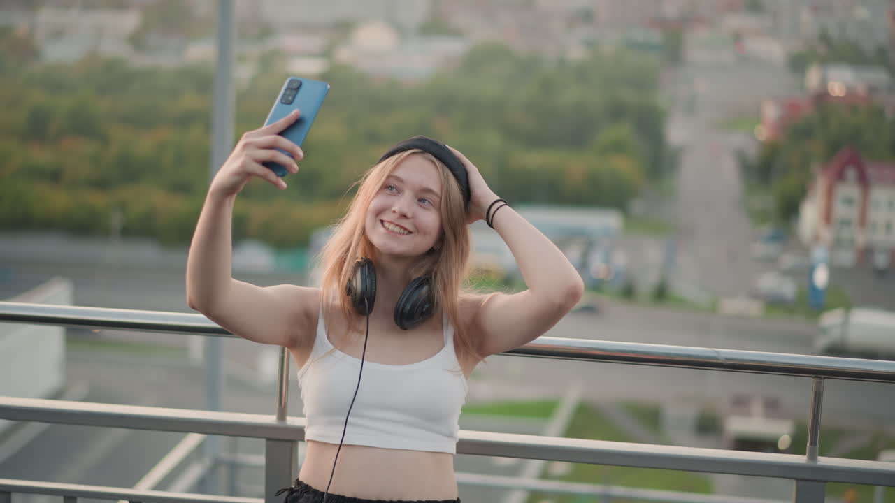 Selfie musicale su un tetto con cuffie, giovane creator registra clip di vlogging spensierati su smartphone, fiume e vista sulla città sullo sfondo, luce dorata della sera, gesti espressivi e umore allegro