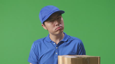 primer plano de un cansado mensajero masculino asiático en uniforme azul sacudiendo la cabeza, tocando su cara y estirándose mientras entrega una caja de cartón en el fondo de la pantalla verde en el estudio