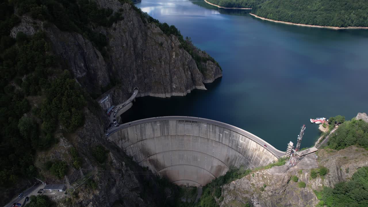 Aerial pullback shot revealing Vidraru Dam and lake in Romania. Blue lake, dramatic curves, and green mountain backdrop in 4K.