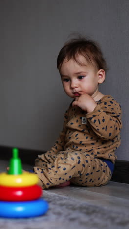 charming kid sitting at the wall in the spacious room holding a finger in his mouth. baby boy looks attentively on the toys in front of him. vertical video