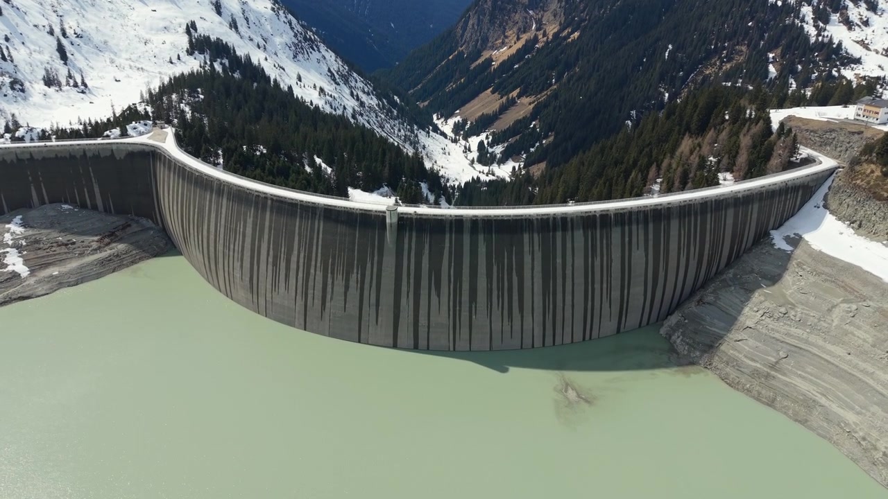 Curved Concrete Dam Holding Pale Green Reservoir In Winter Mountain Valley Near Silvapark In Galtur, Austria. drone tilt-down shot