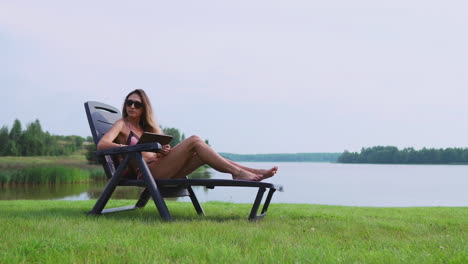 beautiful brunette in a swimsuit lying on a sun lounger on the lake smiling working remotely via the internet in the business sphere. the concept of country life