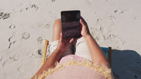 mixed race senior woman sitting on sunbed and using a smartphone at the beach