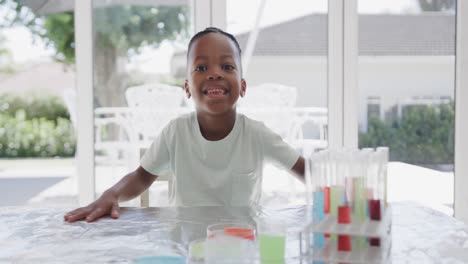 Portrait-of-african-american-boy-sitting-at-table-with-test-tubes-and-smiling,-in-slow-motion