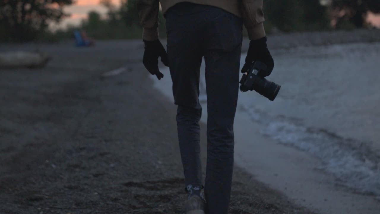 Photographer Holding Camera Walking On The Beach Shore With Ocean Waves And Seagull On The Background. - low-level shot