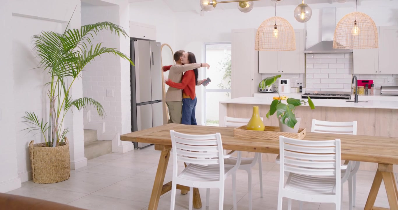 Casal reagindo à entrada da mulher, fazendo o homem encerrar a ligação para cumprimentá-la, movendo-se para a ilha da cozinha