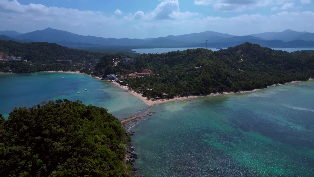 Depeldet Island's lush green landmass connecting to a white sand beach and coastal development, surrounded by clear blue waters. Tremendous aerial view ascending raise up drone panorama