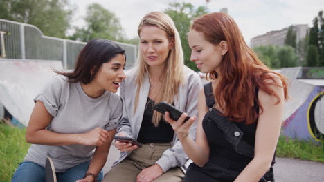 Tres-Amigas-Mirando-El-Teléfono-Móvil-En-El-Parque-De-Patinaje-Urbano-Y-Riendo