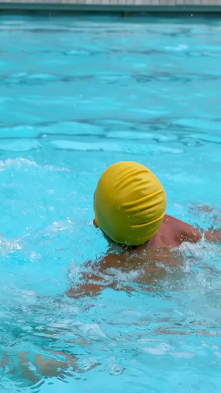 Video vertical: Cámara comenzando a filmar a un niño nadando y practicando brazadas en la piscina, con gorro de natación amarillo.