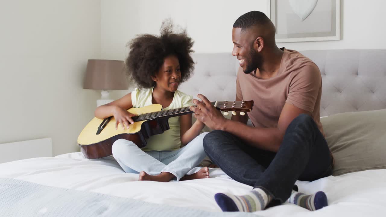 Padre afroamericano e sua figlia seduti sul letto che suonano la chitarra insieme.