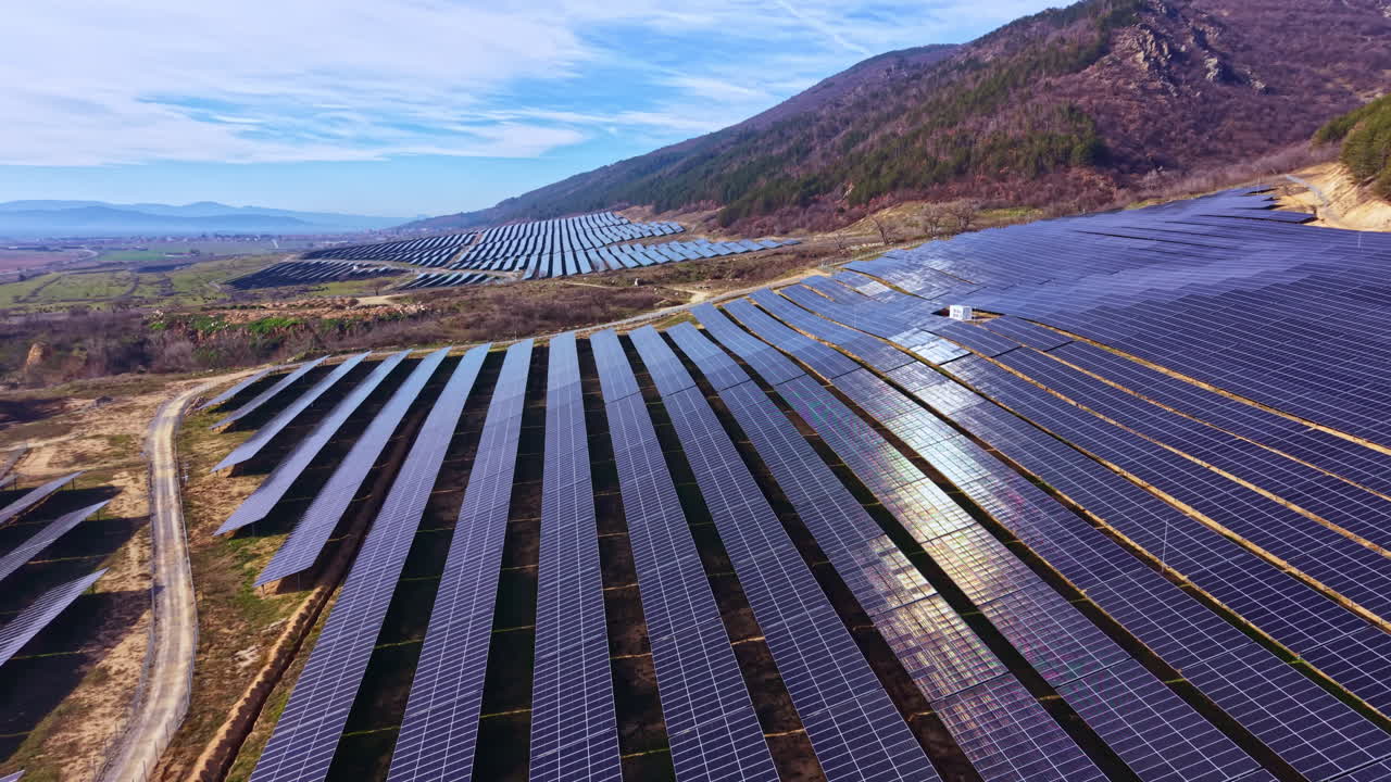 Solpaneler dækker jord på en bjergskråning med klar himmel og marker nedenfor på et energiproduktionssted om dagen
