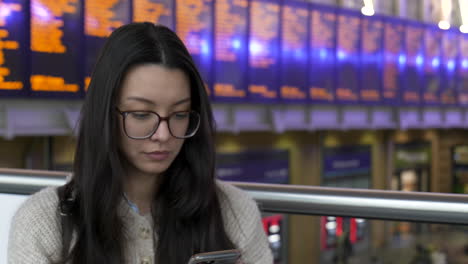 young woman looking at her phone while waiting for her train, station departure boards behind her