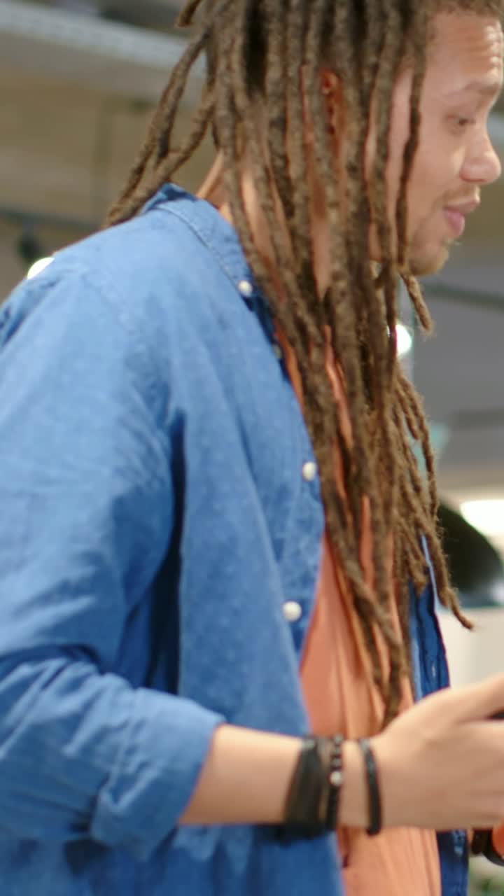 Video verticale: Uomo con dreadlock che si gira e ride al caffè, tiene un telefono, con braccialetti, e indossa un top blu