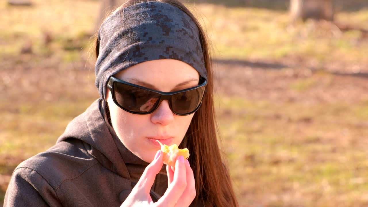 una joven morena con gafas de sol come mandarina sentada en el banco del parque. cara de primer plano.