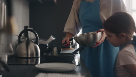 curious little girl is viewing how mother is cooking pancakes pouring dough on frying pan