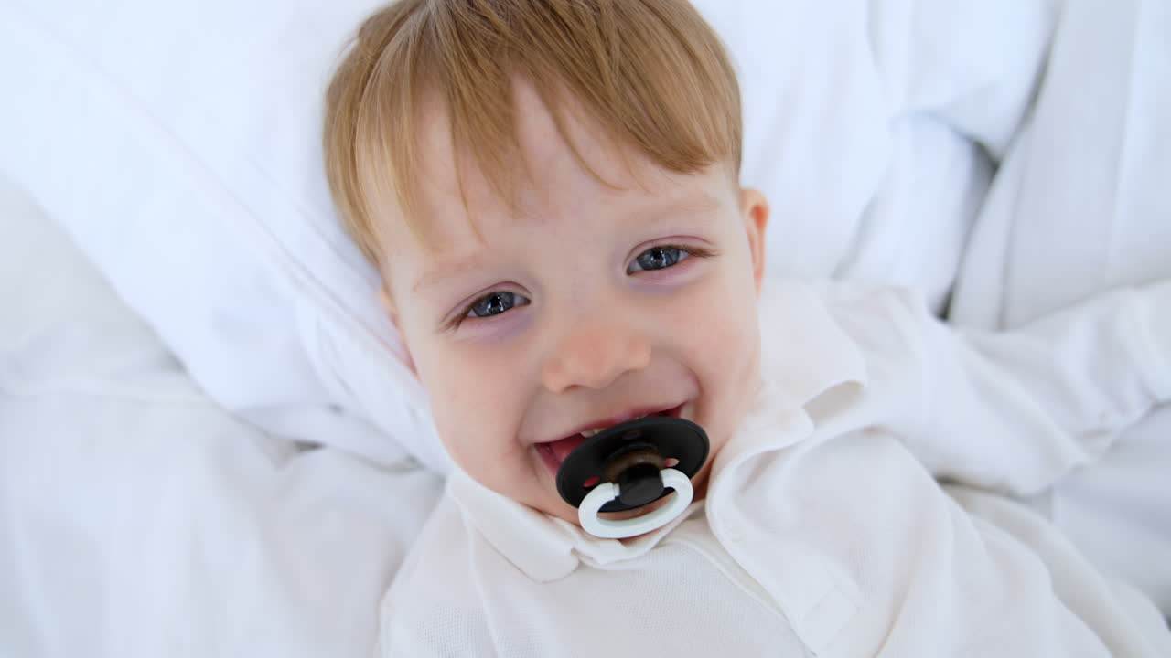 Happy grey-eyed Caucasian baby boy with pacifier in mouth smiling adorably. Cute baby boy lies relaxed on the bed. Close up.