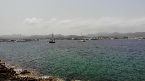 Beautiful-aerial-view-of-limestone-beach-in-Ibiza,-Sant-Antoni-towards-yachts-swaying-in-crystal-clear-emerald-blue-sea-on-a-warm-sunny-day-with-mountains-in-background-in-Spain