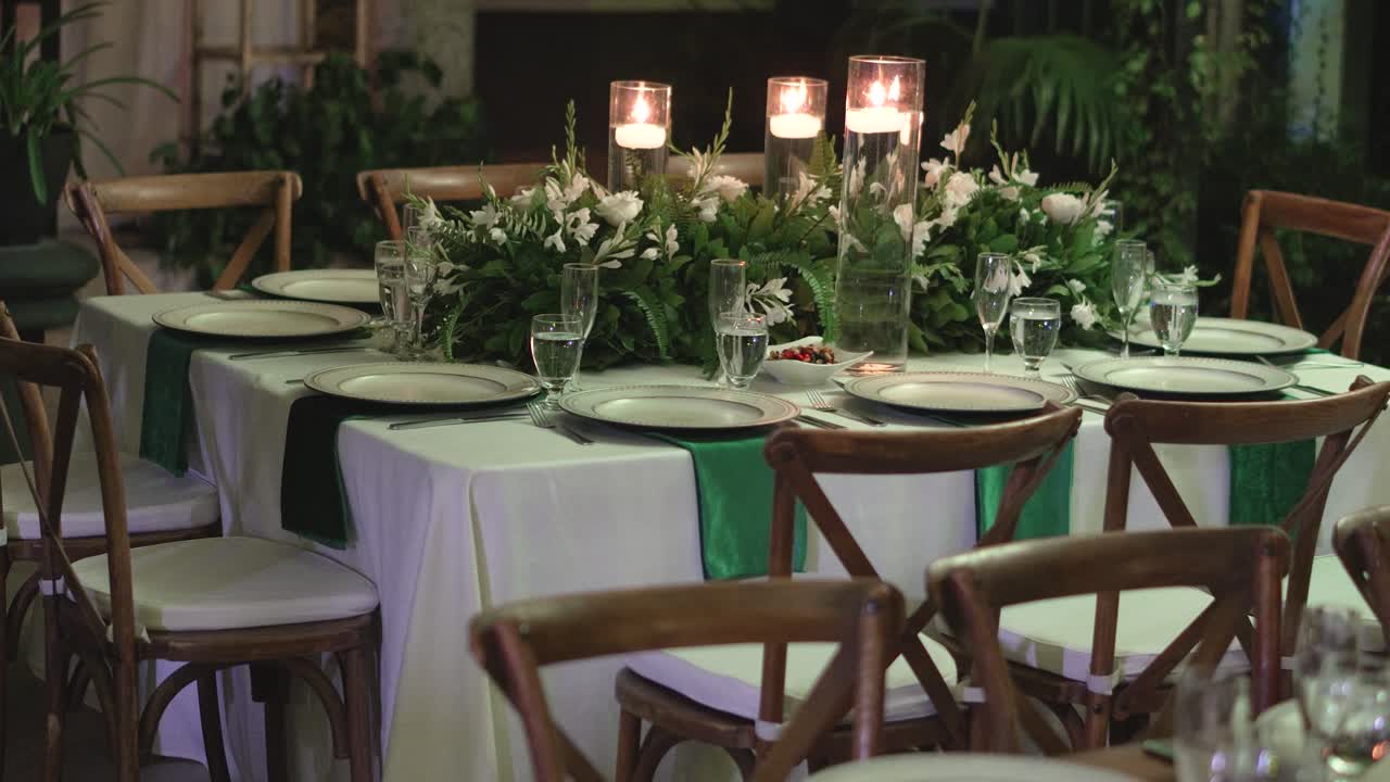 mesa decorada para una boda con un mantel blanco, sillas de madera, artículos de vidrio y piezas centrales con follaje natural