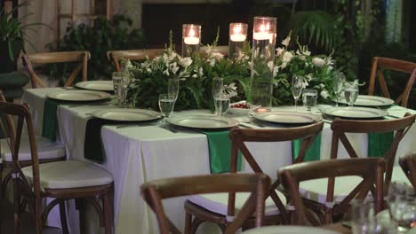 table decorated for a wedding with a white tablecloth, wooden chairs, glassware and centerpieces with natural foliage